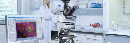 Biowissenschaftler im Laborkittel betrachtet eine Probe mit einem Fluoreszenzbild auf einem Computerbildschirm neben einem Mikroskop im Vordergrund