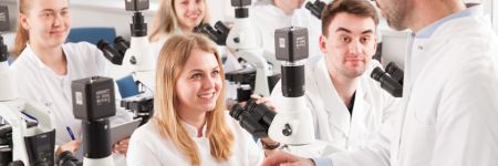 Studierende in Labormänteln in einem Unterrichtsraum für Mikroskopie mit mehreren aufrechten Mikroskopen, die mit Digitalkameras ausgerüstet sind. Der Dozent hält ein Tablet mit dem vergrößerten Bild einer Probe auf dem Bildschirm in den Händen.